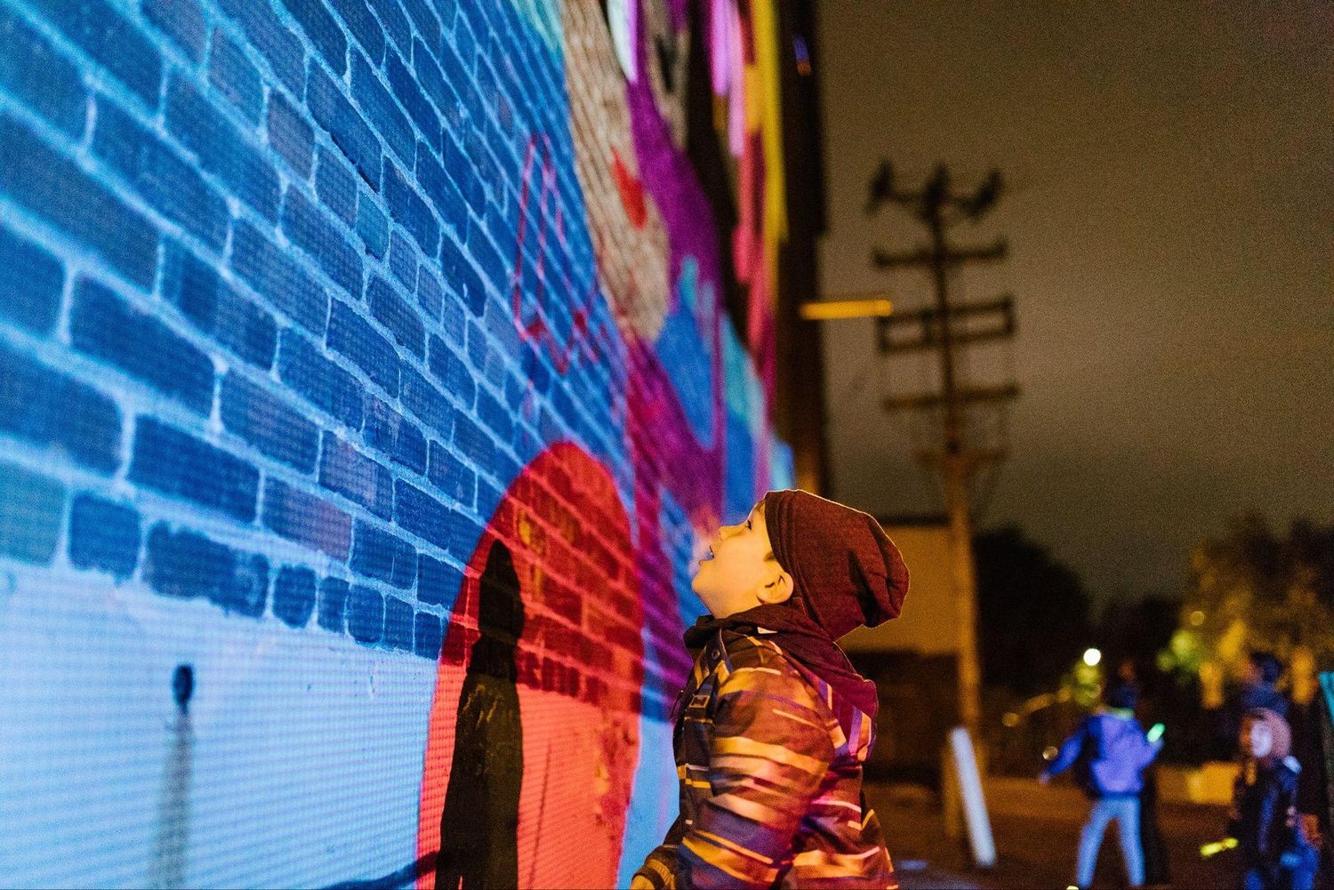 Projection mapping colorée sur un mur de briques, dans la rue, la nuit, devant des passants.
